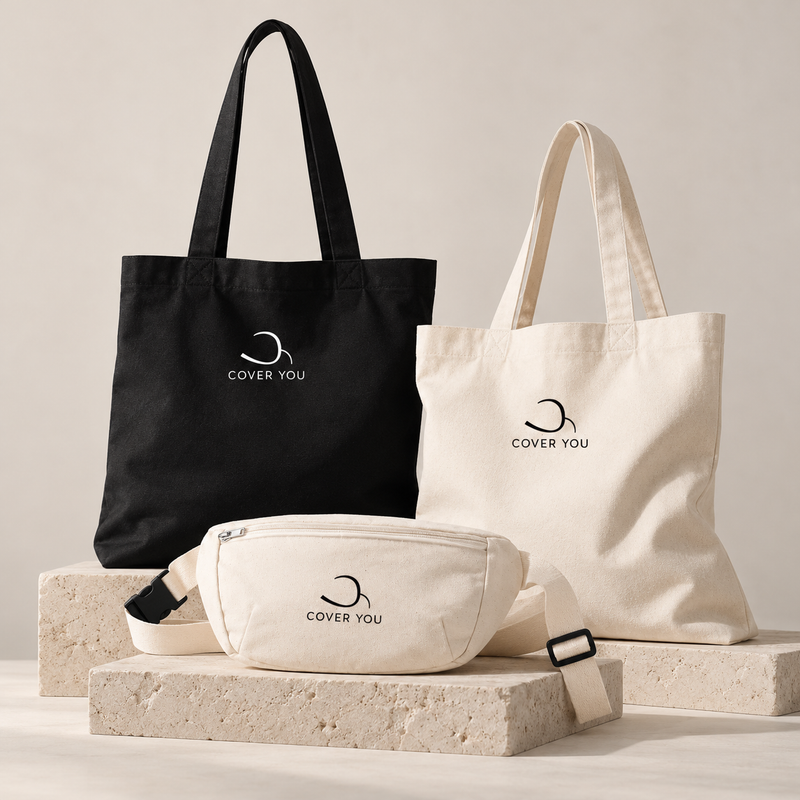 Deux tote bags COVER YOU, noir et écru, et un sac banane écru en coton recyclé, posés sur des blocs de pierre