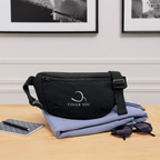 Sac banane COVER YOU noir posé sur une chemise pliée, près d'un carnet et de lunettes de soleil