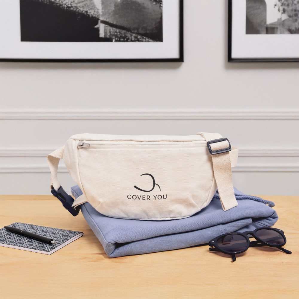 Sac banane COVER YOU naturel posé sur une chemise pliée, près d'un carnet et de lunettes de soleil